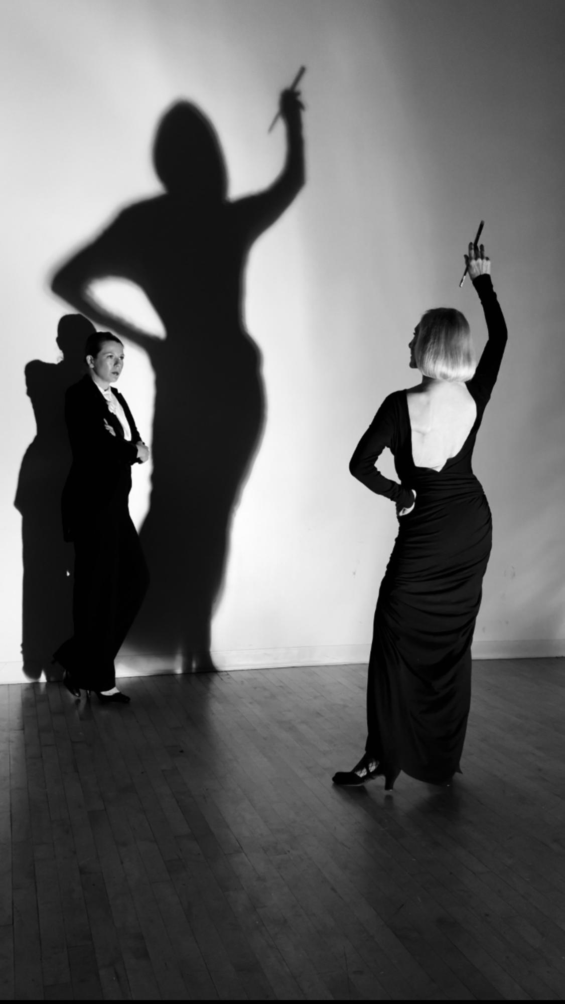 Madame C in a tuxedo and Madame K in a gown holding a paintbrush, her shadow cast large on the studio wall