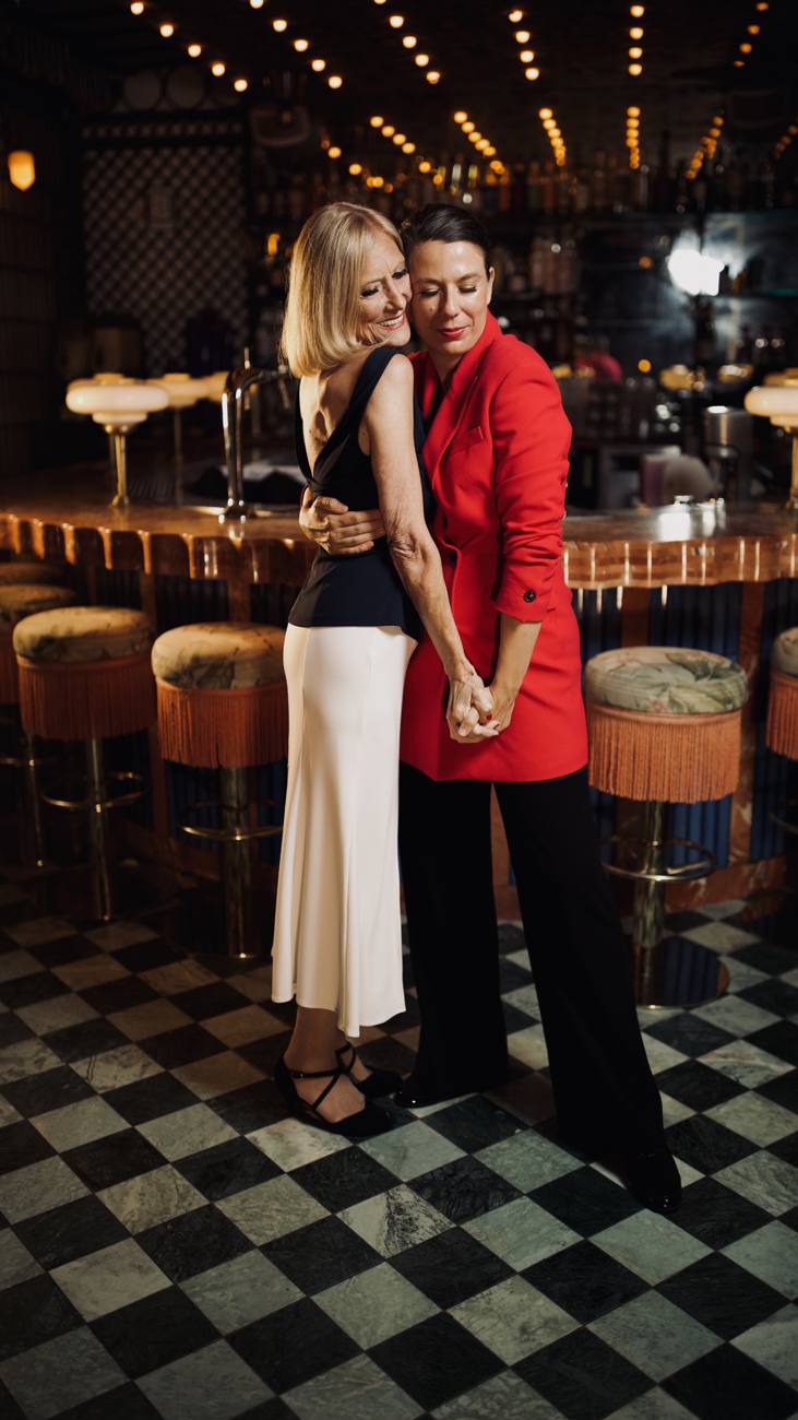 Madame C and Madame K holding hands in an embrace at a bar