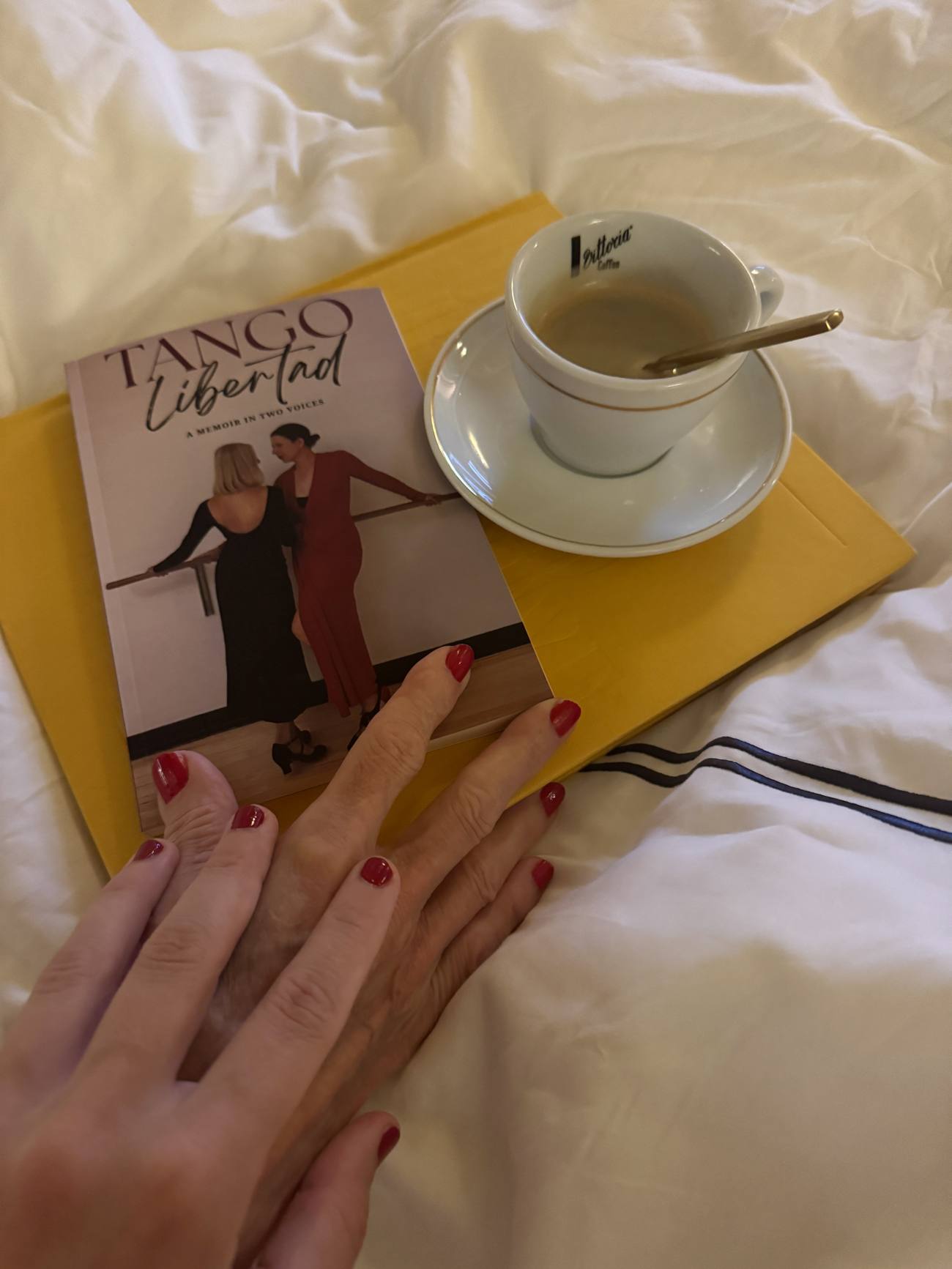 Two hands with red nails resting together beside the memoir and a cup of coffee on a bed